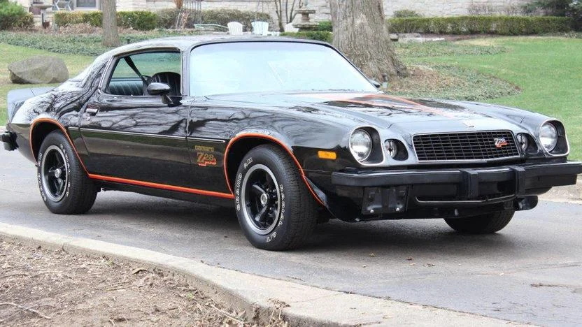 A black classic sports car parked on a street, featuring a sleek design with red accents and a "Z28" emblem on the side. It has a muscular body and sporty wheels, surrounded by a natural setting with trees and grass in the background.