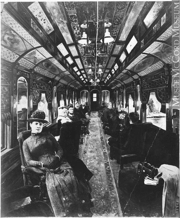 Black and white photo of a vintage train car interior with ornate ceilings and patterned carpet. Passengers in period clothing sit on upholstered seats, some looking towards the camera. Large windows line the sides, offering a view of the landscape.