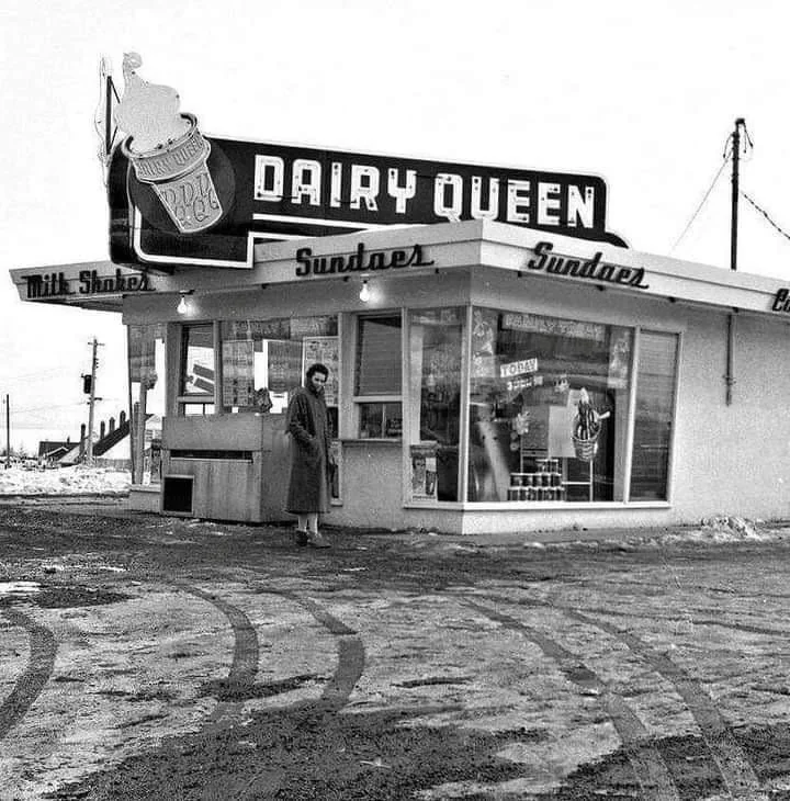 Black and white photo of a vintage Dairy Queen store with a large sign and awning displaying "Sundaes" and "Milk Shakes." A person in a coat stands near the entrance. The ground is snowy, and tire tracks are visible.