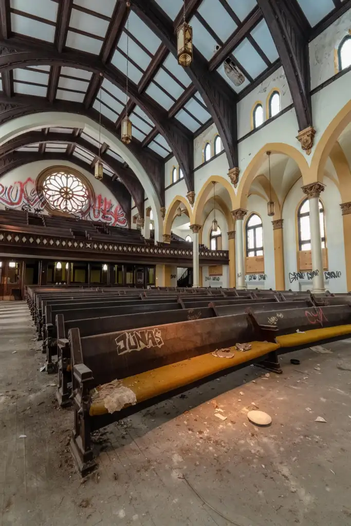 A large, abandoned church interior with empty wooden pews covered in graffiti. The high ceiling features exposed beams and hanging lights. The floor has debris, and the walls include arched windows and a large decorative stained glass window.