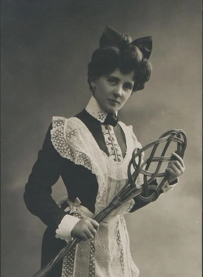 A woman in vintage attire holds a woven carpet beater. She wears a dark dress with lace detailing and a large bow in her hair. The image is in black and white, giving it an antique feel.