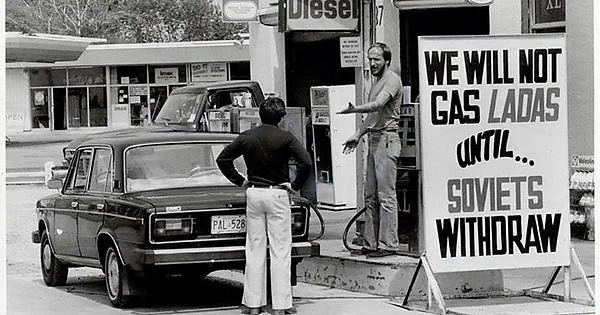 Black and white photo of a man standing at a petrol station with a sign that reads, "WE WILL NOT GAS LADAS UNTIL... SOVIETS WITHDRAW." A Lada car is parked at the station, and another man stands nearby. A shop is visible in the background.