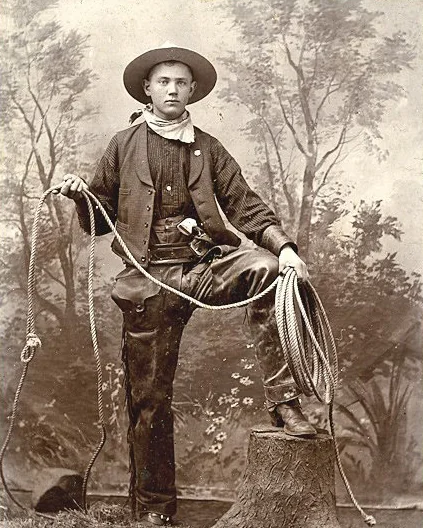 Vintage photo of a cowboy standing with one foot on a tree stump, wearing a wide-brimmed hat, vest, and chaps. He's holding a rope and has a gun holstered at his waist. The backdrop features painted trees and foliage.