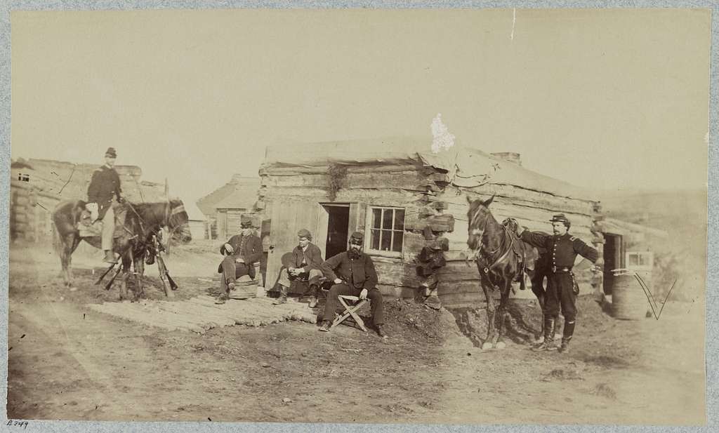 A vintage photo shows several men in military uniforms outside a log cabin in a barren landscape. Two are on horseback, while others stand or sit nearby. A few rifles are visible, and there’s another small cabin in the background.