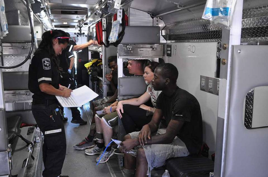 A paramedic checks paperwork inside an ambulance while several people sit on benches along the sides. The interior is equipped with medical supplies and equipment. One person holds a clipboard, and medical bags are visible above.