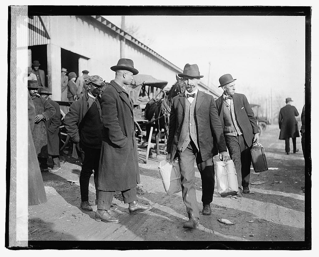 Black and white photo of men in coats and hats walking outside a building. Two men carry bags or containers. The scene includes a crowd in the background, possibly at a market or station. The ground is unpaved.