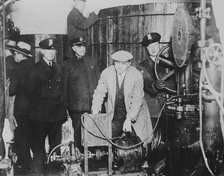 A historical black-and-white photo showing a group of men in early 20th-century clothing and hats. Most are wearing uniforms, while one operates industrial machinery with large pipes and wheels in a ship or factory setting.