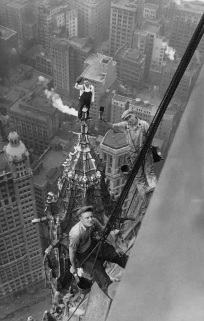 Black and white photo of three construction workers high above a city skyline. Two workers are standing on the spire of a tall building, and one is sitting and holding a cable. Skyscrapers and smoke-filled chimneys are visible in the background.