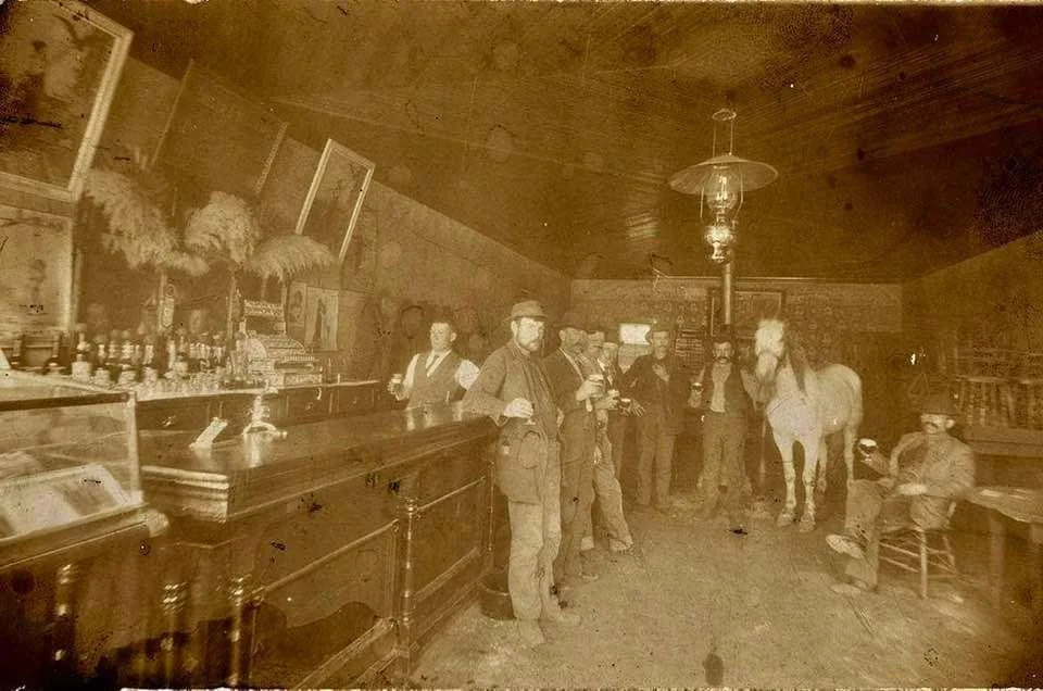 Sepia-toned image of a vintage bar scene with several men in period attire standing around a wooden bar. An oil lamp hangs above. A horse stands inside near the men. The room features framed pictures and ornate decor.