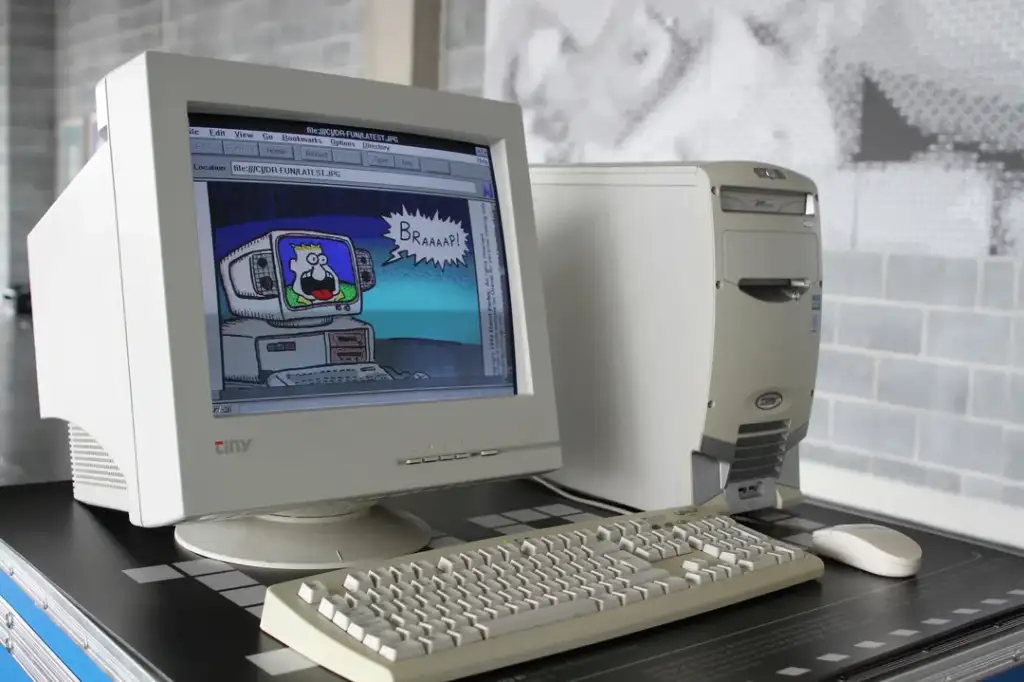 A vintage desktop computer setup with a CRT monitor, keyboard, and mouse sits on a desk. The monitor displays a cartoon image of a computer with the text "BRAAAP!" in a speech bubble. The computer tower is off-white and rectangular.