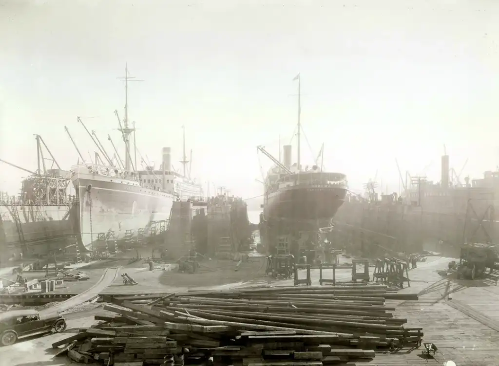 A vintage black-and-white photo depicting a bustling dry dock with two large ships being repaired. Wooden beams and materials are scattered across the dock, while numerous workers are engaged in activities around the ships.