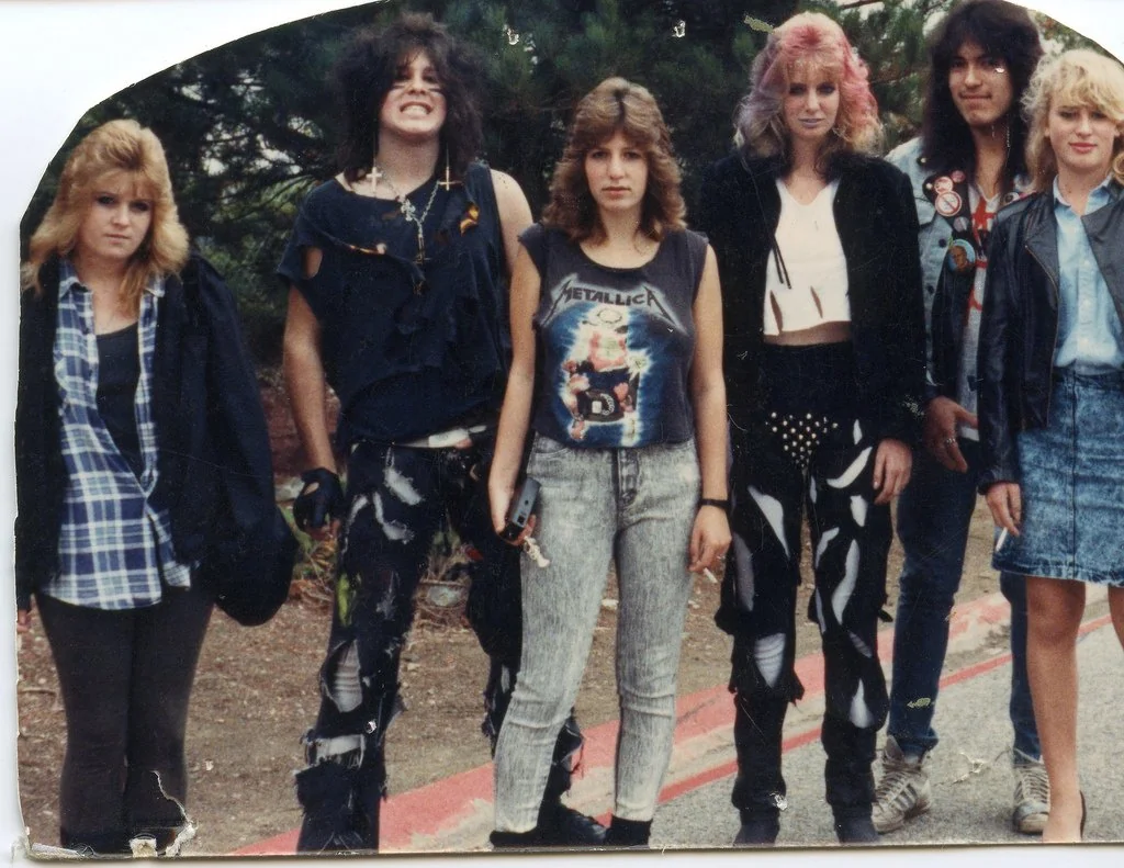 Six people stand outdoors in a casual group. They are dressed in 1980s punk and rock styles, featuring denim, leather jackets, and band t-shirts. Some have teased or dyed hair. The background includes greenery and a pathway.