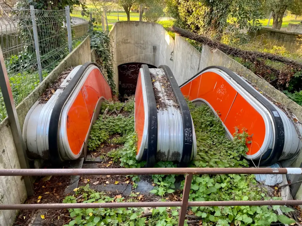 Overgrown outdoor escalators lead into an underground passage. The metal steps and sides are coated with graffiti and leaves. Vegetation grows abundantly, and trees and a fence are visible in the background.