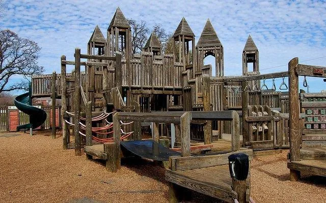 Wooden playground structure featuring towers, bridges, slides, and climbing elements, set on a mulch-covered ground with a clear blue sky above. Trees are visible in the background.