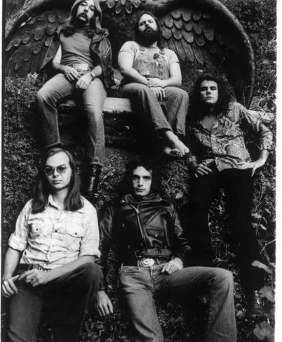 Five men posing in front of a stone angel statue. Two are sitting on the statue's base, and three are seated below. They're dressed in 1970s clothing, including jeans and leather. The image is in black and white.