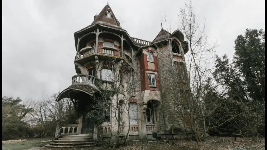 A vintage, multi-story house with turrets and a steep roof stands surrounded by overgrown bushes and bare trees. The building features a mix of stone and brick, with ornate balconies and arched windows, creating a mysterious, gothic atmosphere.