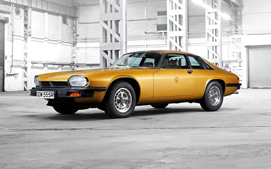 A vintage gold sports car is parked in an industrial warehouse with a concrete floor. The car has a sleek design with a coupe body and chrome details. The white walls and metal beams create a stark, minimalist background.