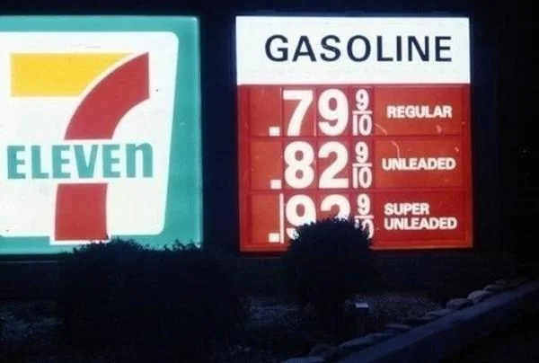 A vintage gas station sign displays gasoline prices: Regular at 79.9 cents, Unleaded at 82.9 cents, and Super Unleaded at 92.9 cents. The 7-Eleven logo is visible next to the sign. Bushes are in front of the signs.