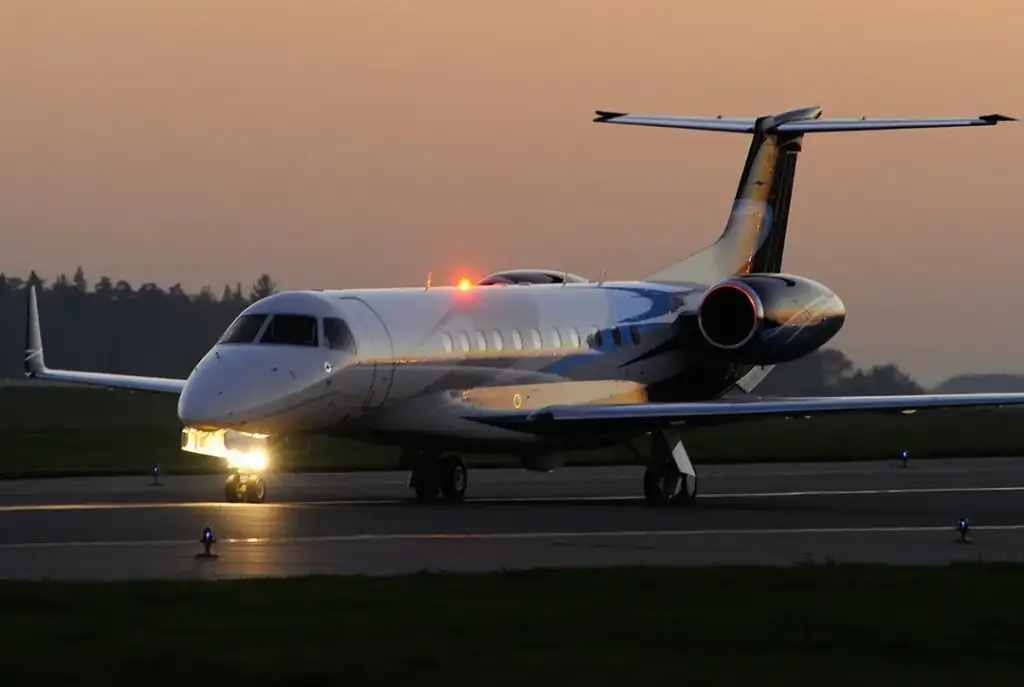 A small jet is taxiing on a runway during sunset, with lights on. The sky is a gradient of orange and blue, and trees are visible in the background.