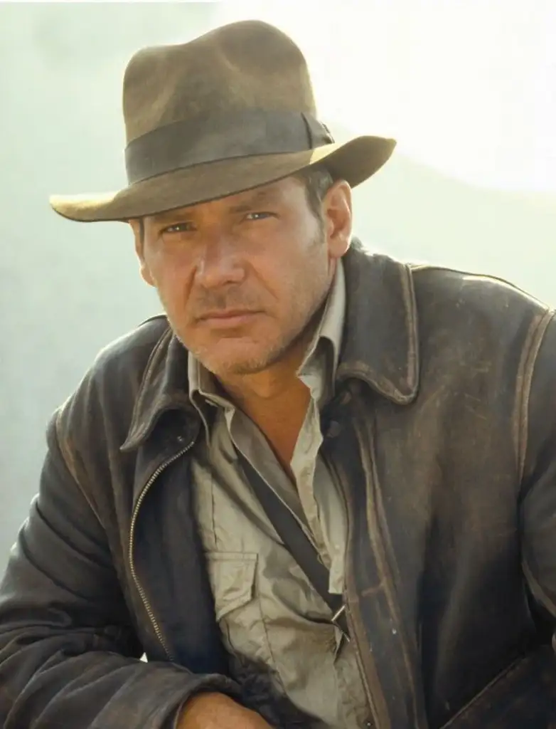 A person wearing a brown fedora and a rugged leather jacket over a beige shirt. They have a serious expression and are outdoors with a neutral background.