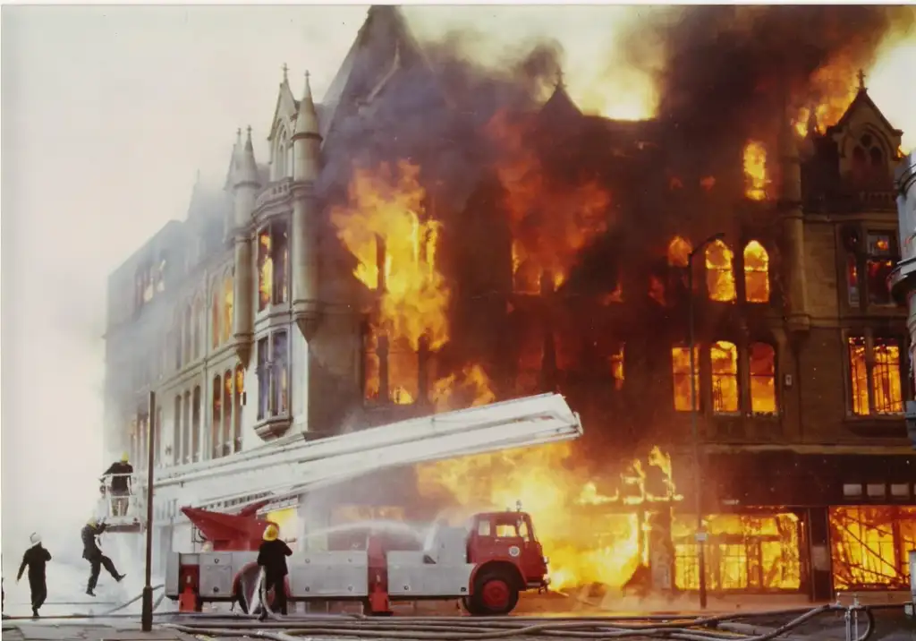 A historic building is engulfed in flames, with large fire visible through windows. Firefighters work at the scene, using hoses and an aerial ladder from a fire truck. The sky is smoky, and debris is scattered on the ground.