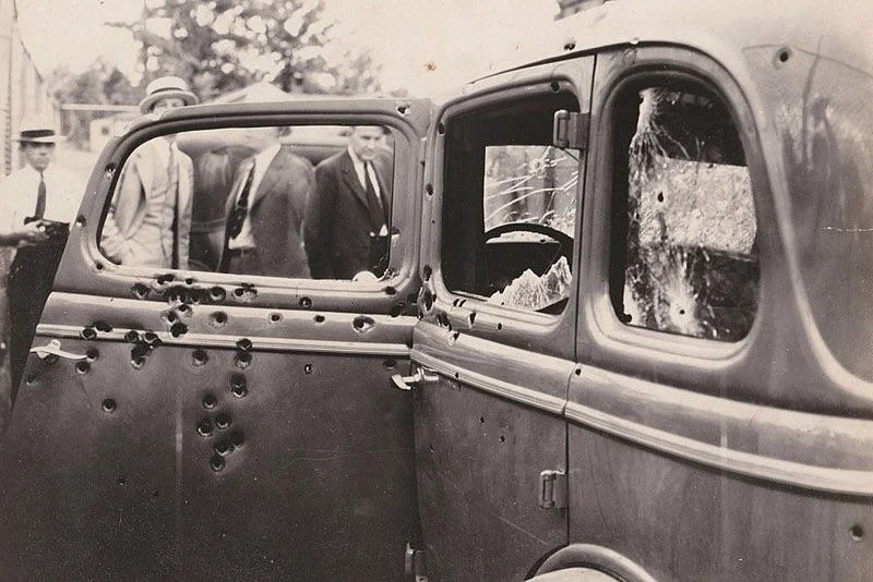 A vintage black-and-white photograph shows an old car riddled with bullet holes and broken windows. Several men in suits and hats stand in the background, observing the scene. Trees are visible in the distance.