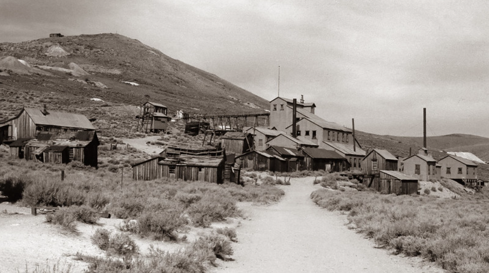 A sepia-toned image of an abandoned western town showing a collection of wooden buildings, dirt paths, and surrounding scrubby vegetation. A barren hill rises in the background under a cloudy sky.