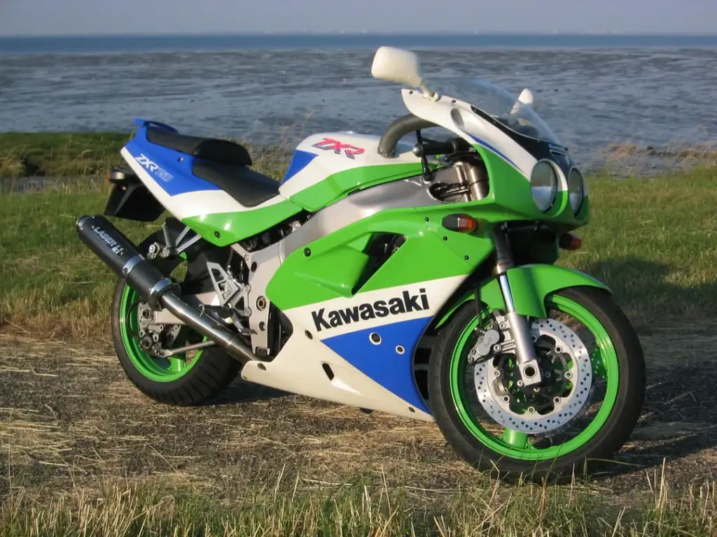 A green, white, and blue Kawasaki sport motorcycle is parked on a grassy area beside a body of water. The bike has a sleek design with "ZX" and "Kawasaki" markings, and the background shows a distant, serene seascape.