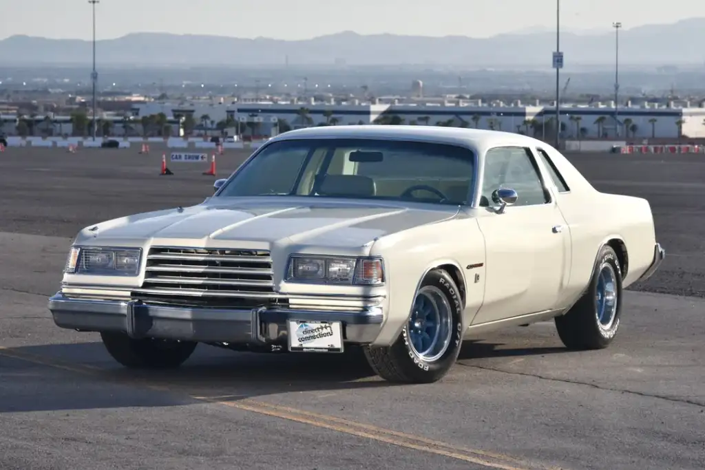 A white vintage car, likely from the late 1970s to early 1980s, is parked in an expansive outdoor area. It features a two-door design and chrome detailing. In the background, there are mountains and a cityscape under a clear sky.