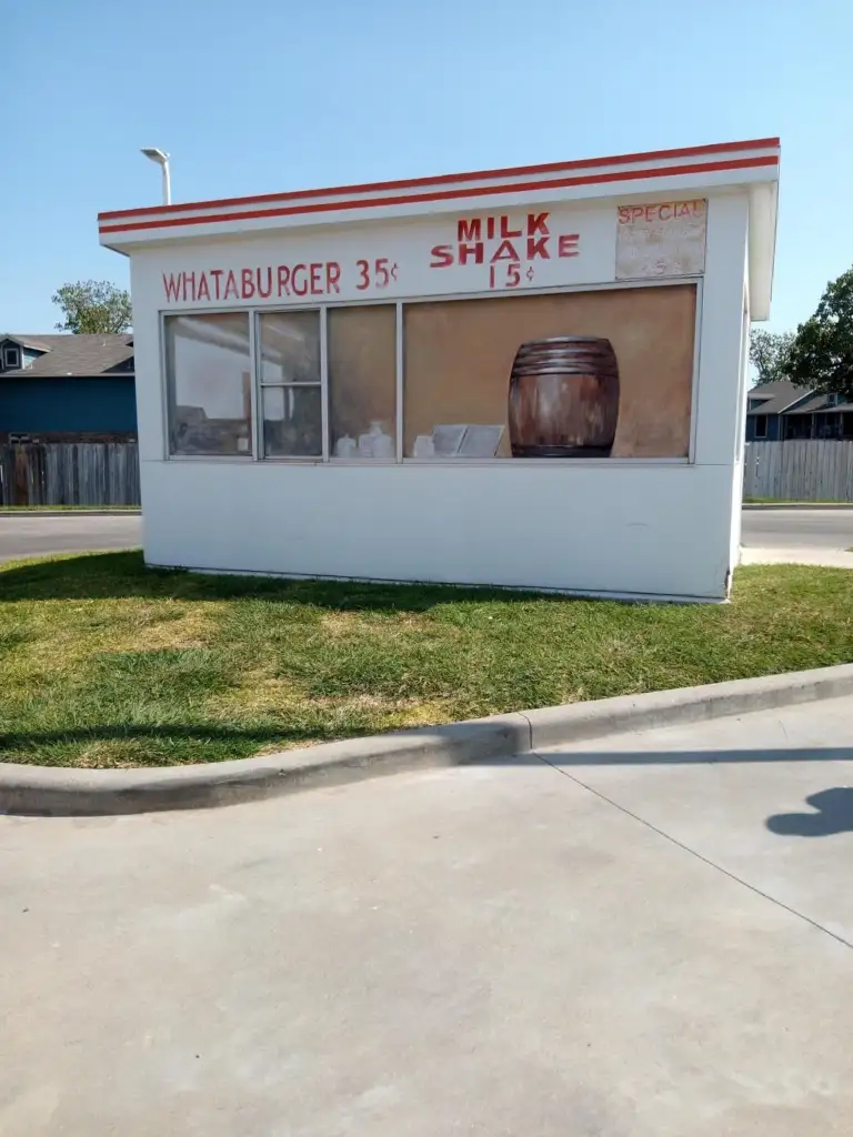 Small vintage diner with signs advertising "Whataburger 35¢" and "Milk Shake 15¢" on the window. The building is white with red trim, set on a grassy area with a wooden fence and houses in the background.