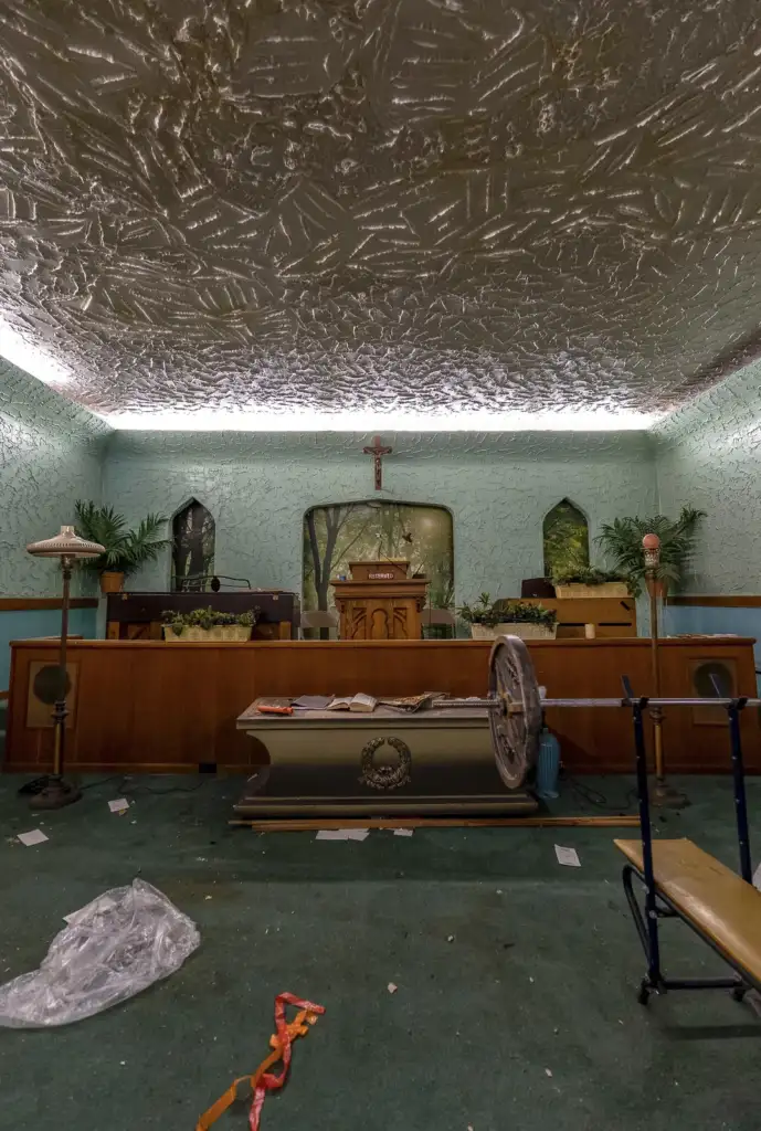 An abandoned church interior with a dusty altar, wooden pews, and a decorative ceiling. The altar features plants and a cross. The floor is cluttered with debris, including a broken wheelchair and scattered papers. Dim lighting creates a somber atmosphere.