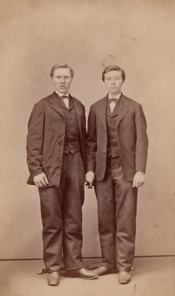 Two young men stand close together, facing forward in a vintage sepia-toned photograph. Both wear dark suits with vests, white shirts, and ties. They have short hair and maintain serious expressions against a plain backdrop.