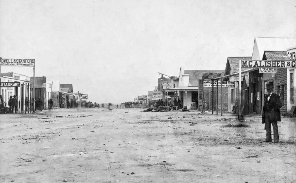 A black and white photo of a wide, deserted dirt street in a historic Western town. Wood and brick buildings line both sides, featuring signs for businesses like a restaurant and store. A few people and a horse-drawn cart are visible in the distance.