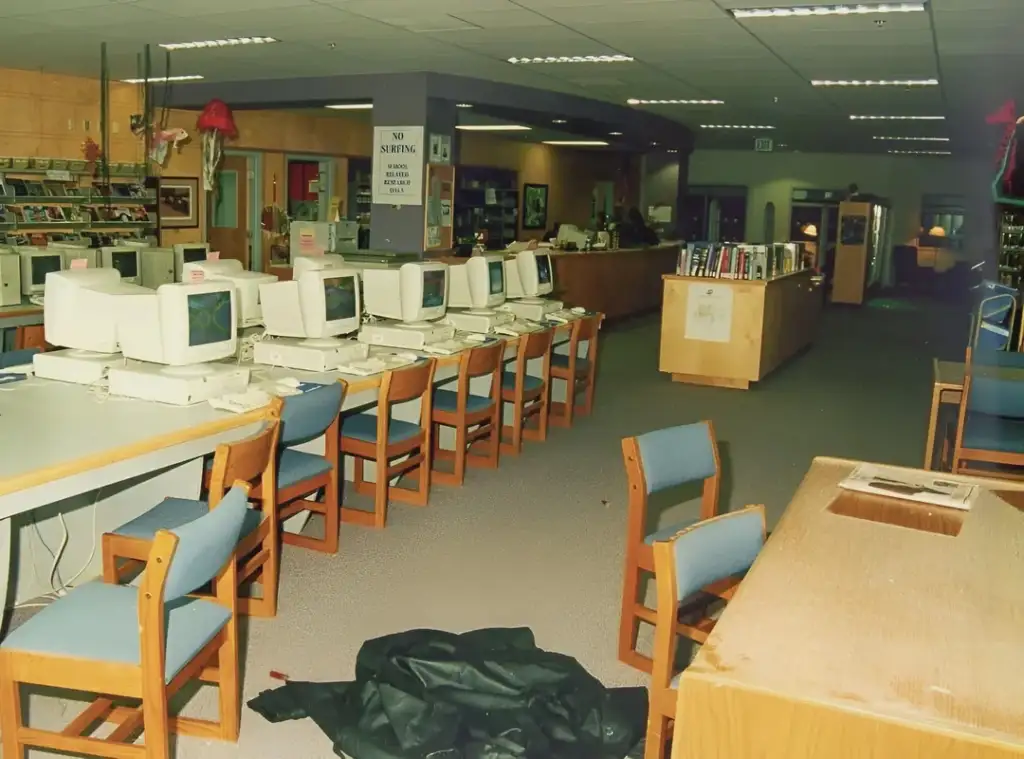 A library with a row of vintage computers on a desk with wooden chairs in front. Shelves filled with books are in the background. A "No Smoking" sign is visible on a column. Carpeted flooring and overhead lights illuminate the space.