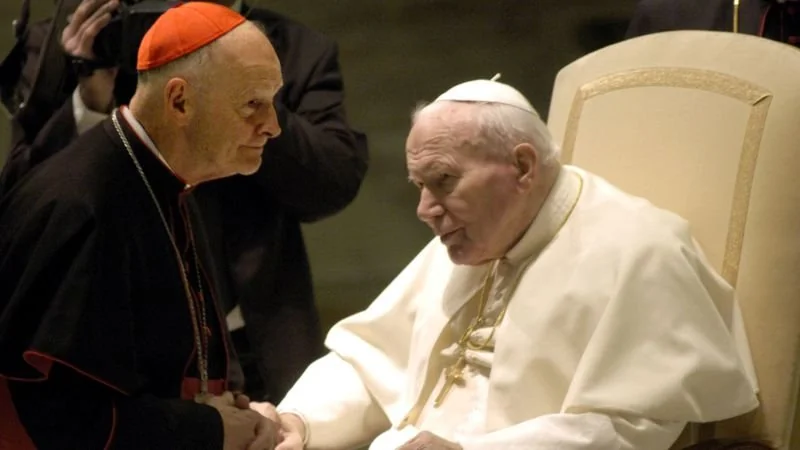 A cardinal in a red cap and black robe leans towards an elderly pope sitting in a white robe and skullcap on an ornate chair. They are engaged in conversation, with the pope holding the cardinal's hand.