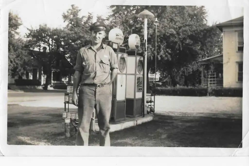 A black and white photo shows a man in a uniform standing in front of vintage gas pumps. He wears a cap and has one hand in his pocket. The background features trees and a building with visible windows.