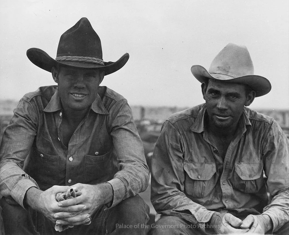 Two men wearing wide-brimmed hats sit side by side outdoors. They both wear button-up shirts and have relaxed expressions. The background is out of focus, suggesting an open landscape or rural setting.