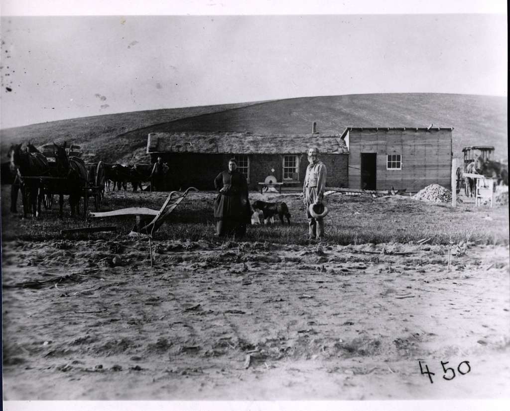 Black and white photo of two people standing in a field in front of a rustic house and barn. A dog sits beside them. Horses and a plow are on the left. A hill rises in the background under a cloudy sky.