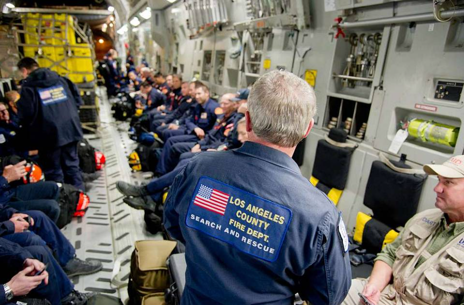 A group of uniformed personnel from the Los Angeles County Fire Dept. Search and Rescue team are sitting inside a large aircraft. The interior is filled with gear and equipment. One member, with his back to the camera, wears a jacket with the team's emblem.