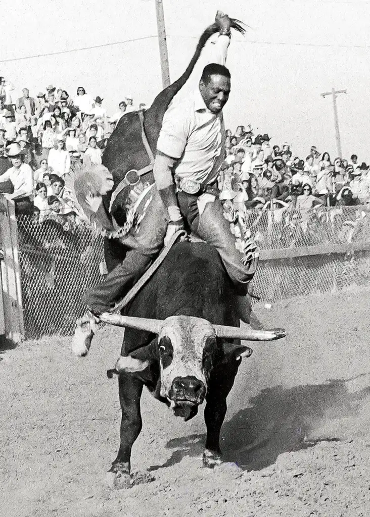 A man rides a bull during a rodeo event, holding onto the bull with one hand and raising the other. The bull is in mid-leap, exhibiting its fierce energy. Spectators watch behind a fence, creating an intense and dynamic scene.