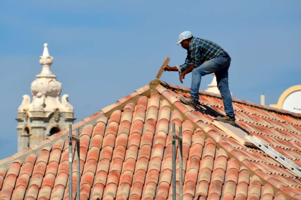 Person in a plaid shirt and hat working on a tiled roof against a clear blue sky. A decorative building structure is visible in the background.