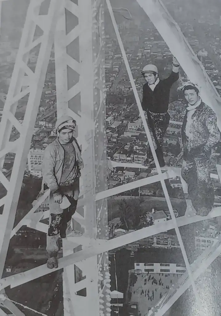 Three construction workers stand on the steel frame of a tall structure overlooking a cityscape. They are wearing protective gear, including helmets. The city is visible in the background, showing buildings and roads below.