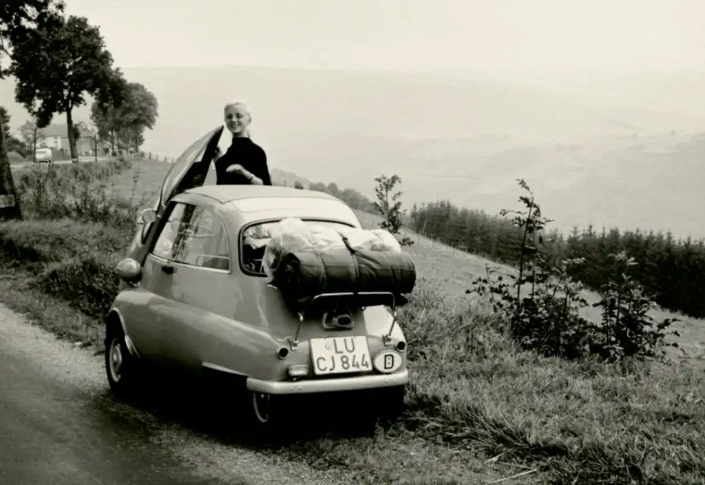 A smiling person stands next to a small vintage car with a large bag strapped to the back. The car is parked on a road with scenic hills and trees in the background. The image is in black and white.