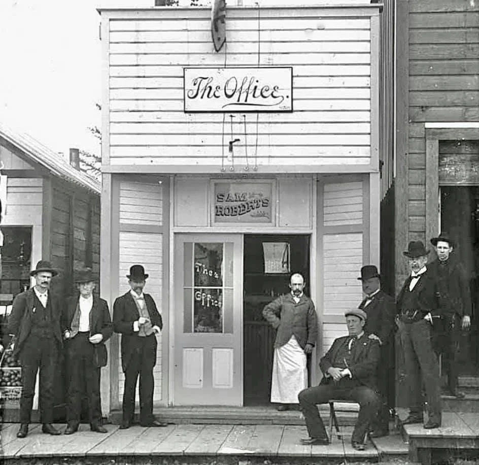 Black and white photo of eight men in 19th-century attire standing and sitting outside a wooden building labeled "The Office." The storefront has a sign for "Sam's Rooms," and the men appear relaxed and social.