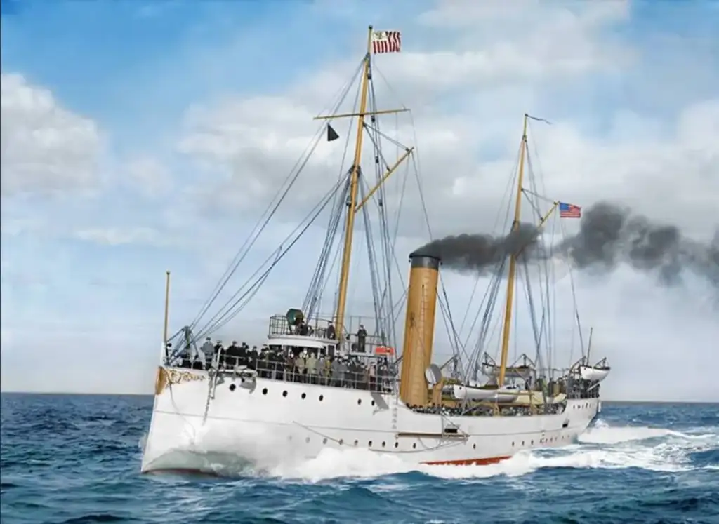 A vintage steamship sailing on a calm ocean under a clear sky. The ship has a tall smokestack emitting black smoke and is adorned with U.S. flags. The hull is white with visible lifeboats and passengers on deck.