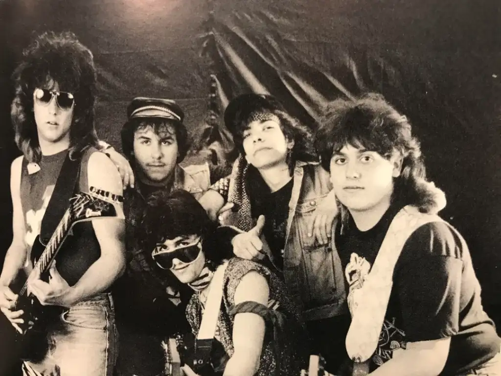 A black and white photo of a five-member band. Four are standing and one is seated, all wearing edgy rock-style clothing. Two members hold guitars. Their expressions convey confidence and attitude, and they are positioned against a draped backdrop.