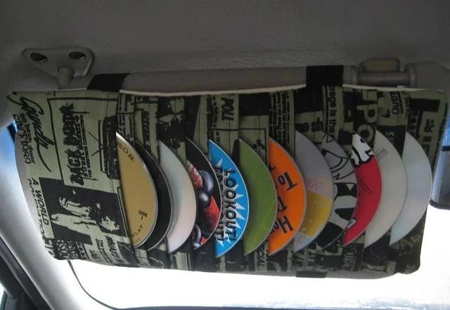 A car visor organizer holding various CDs with colorful labels. The organizer has a vintage newspaper-themed fabric. It is attached to the car's visor, visible from the driver's side.