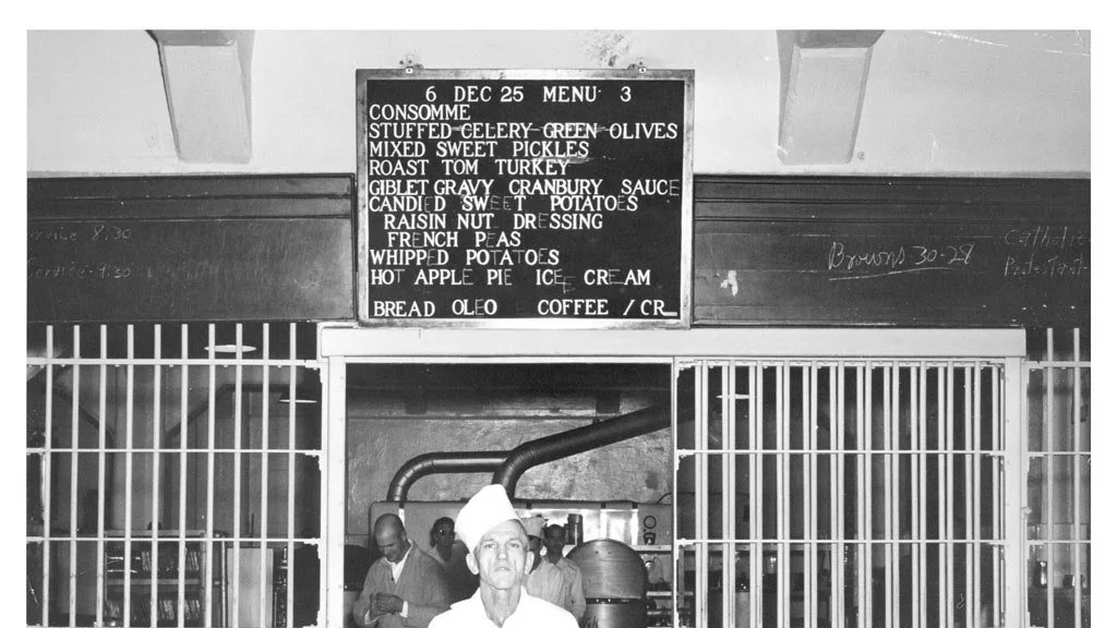 A black-and-white photo shows a chef standing in front of a kitchen with a menu board overhead. The menu for December 6, 1925, includes consommé, roast tom turkey, French peas, whipped potatoes, and hot apple pie. Kitchen staff work in the background.