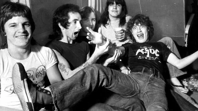 A black-and-white photo of five people posing playfully. One person on the far right is being held horizontally by two others, smiling widely. The group appears cheerful and relaxed. The person being held wears an AC/DC shirt.