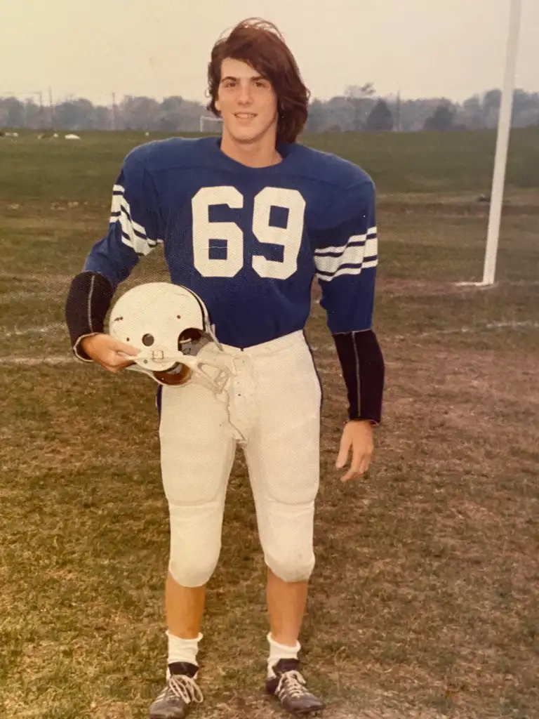 A young football player stands on a grass field wearing a blue jersey with the number 69 and white pants. He holds a white helmet under one arm. The background shows an open field and goalpost.